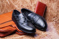 A pair of black leather loafers with tassel detailing is placed on a brown leather briefcase. A red leather wallet is set against a marble-textured background, creating a luxurious and elegant arrangement.