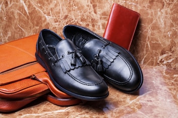 A pair of black leather loafers with tassel detailing is placed on a brown leather briefcase. A red leather wallet is set against a marble-textured background, creating a luxurious and elegant arrangement.