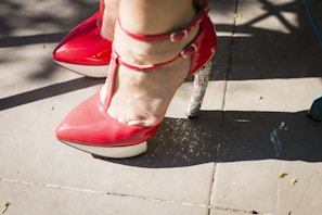 A close-up of red high-heeled shoes with ankle straps, one with a glittering silver heel. The shoes are on a paved outdoor surface with some scattered shadows and small leaves.