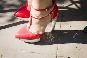 A close-up of red high-heeled shoes with ankle straps, one with a glittering silver heel. The shoes are on a paved outdoor surface with some scattered shadows and small leaves.