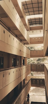 A multi-level atrium with balconies on each floor, featuring a rooftop that allows natural light to enter. The architectural design is modern and minimalistic, with beige walls and small square windows. At the ground level, a person walks through the corridor.