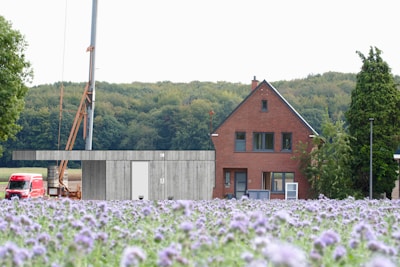 A brick house with a steep roof is set against a background of dense green forest. The foreground features a field of purple flowers. To the left, there is a partially constructed wind turbine with a crane beside it. A red vehicle is parked near the construction area.