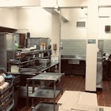 A professional kitchen setting with stainless steel shelves and countertops, featuring an array of kitchen supplies and equipment. The room has tiled flooring and walls, with various silver and gray tones dominating the decor.
