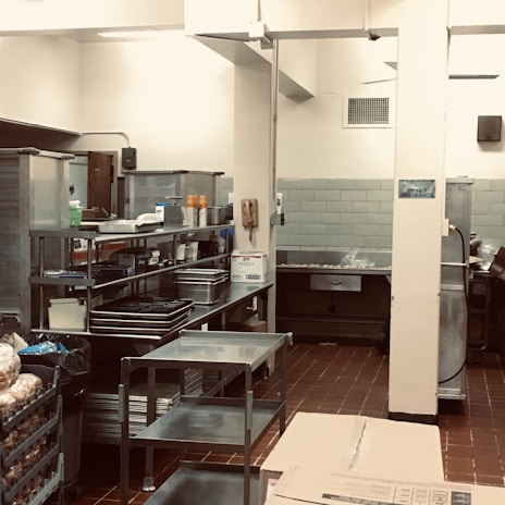 A professional kitchen setting with stainless steel shelves and countertops, featuring an array of kitchen supplies and equipment. The room has tiled flooring and walls, with various silver and gray tones dominating the decor.