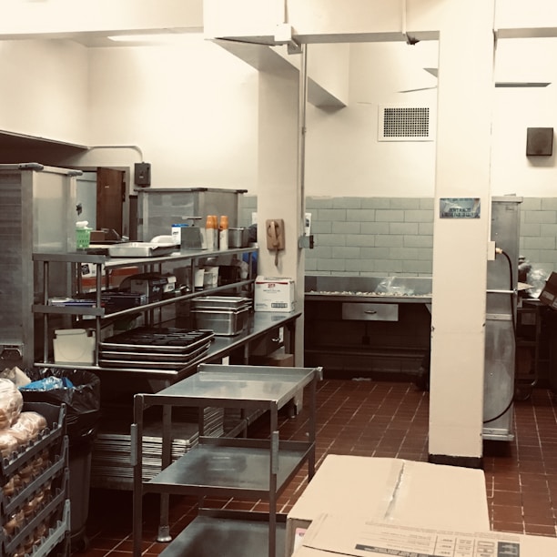 A professional kitchen setting with stainless steel shelves and countertops, featuring an array of kitchen supplies and equipment. The room has tiled flooring and walls, with various silver and gray tones dominating the decor.
