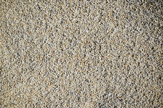A close-up view of a large number of small, roughly textured pebbles or gravel. The pebbles vary in color, predominantly featuring shades of beige, gray, and light brown. The composition creates a uniform, natural pattern with subtle variations in color.