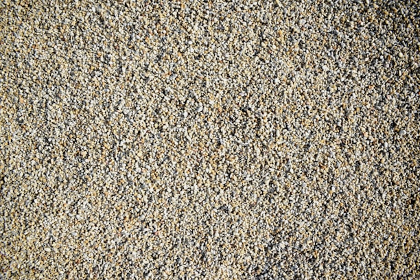A close-up view of a large number of small, roughly textured pebbles or gravel. The pebbles vary in color, predominantly featuring shades of beige, gray, and light brown. The composition creates a uniform, natural pattern with subtle variations in color.