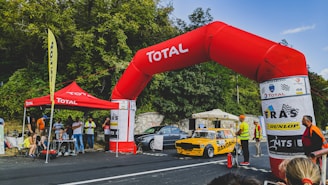 A yellow rally car with the branding Bardahl is positioned under a large red inflatable arch labeled Total. Several people, including officials and spectators, are gathered around, some wearing safety vests. There's a Total tent nearby with people sitting under it, and a Dunlop banner is visible. The background features dense foliage and a clear sky.