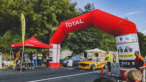 A yellow rally car with the branding Bardahl is positioned under a large red inflatable arch labeled Total. Several people, including officials and spectators, are gathered around, some wearing safety vests. There's a Total tent nearby with people sitting under it, and a Dunlop banner is visible. The background features dense foliage and a clear sky.