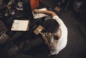 A person is seated at a cluttered workspace, organizing or packaging items. Several clipboards with papers, a notebook, and a variety of tools and boxes are scattered across a wooden table. The individual is wearing a white shirt and appears to be focused on their task.