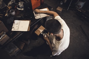 A person is seated at a cluttered workspace, organizing or packaging items. Several clipboards with papers, a notebook, and a variety of tools and boxes are scattered across a wooden table. The individual is wearing a white shirt and appears to be focused on their task.