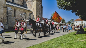 A historical parade with people dressed in traditional uniforms, marching alongside a building with stone architecture. Two individuals on horseback carry an orange flag, followed by others holding wooden poles. Spectators are gathered along the sidewalk, enjoying the sunny day.