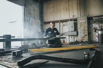 A person wearing a work uniform stands in an industrial workshop. The environment is filled with metal tools and equipment, and there is a faint haze of smoke in the air. High voltage signs and machinery control boxes are visible along the walls.