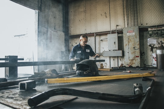 A person wearing a work uniform stands in an industrial workshop. The environment is filled with metal tools and equipment, and there is a faint haze of smoke in the air. High voltage signs and machinery control boxes are visible along the walls.