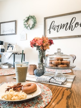 A cozy kitchen scene with a wooden table set with a plate of breakfast food including scrambled eggs, bacon, and a donut. An iced coffee is in a clear glass with a straw. In the center of the table, there is a floral arrangement and a cake stand with donuts under a glass dome. The wall is adorned with farmhouse-style decor, including a wreath and framed signs.
