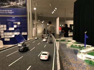 A detailed diorama of a road featuring miniature cars is displayed. The scene extends into an exhibition space where several people can be observed in the background, engaging with various exhibits. The setting includes informational panels and artistic elements, with a focus on transportation infrastructure.