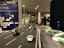 A detailed diorama of a road featuring miniature cars is displayed. The scene extends into an exhibition space where several people can be observed in the background, engaging with various exhibits. The setting includes informational panels and artistic elements, with a focus on transportation infrastructure.