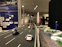 A detailed diorama of a road featuring miniature cars is displayed. The scene extends into an exhibition space where several people can be observed in the background, engaging with various exhibits. The setting includes informational panels and artistic elements, with a focus on transportation infrastructure.