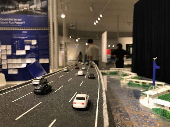 A detailed diorama of a road featuring miniature cars is displayed. The scene extends into an exhibition space where several people can be observed in the background, engaging with various exhibits. The setting includes informational panels and artistic elements, with a focus on transportation infrastructure.