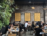 A lively café environment with several people sitting at wooden tables, engaged in conversation or working on laptops. The walls are made of exposed brick and decorated with framed text prints. A vertical plant installation adds greenery to one side. The warm lighting creates a cozy atmosphere.