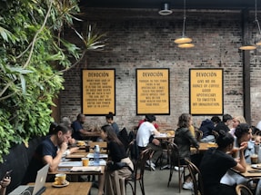 A lively café environment with several people sitting at wooden tables, engaged in conversation or working on laptops. The walls are made of exposed brick and decorated with framed text prints. A vertical plant installation adds greenery to one side. The warm lighting creates a cozy atmosphere.