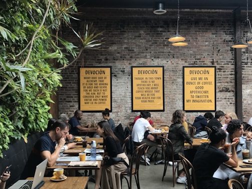A lively café environment with several people sitting at wooden tables, engaged in conversation or working on laptops. The walls are made of exposed brick and decorated with framed text prints. A vertical plant installation adds greenery to one side. The warm lighting creates a cozy atmosphere.