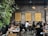 A lively café environment with several people sitting at wooden tables, engaged in conversation or working on laptops. The walls are made of exposed brick and decorated with framed text prints. A vertical plant installation adds greenery to one side. The warm lighting creates a cozy atmosphere.