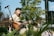 A man smiling while strumming a banjo outdoors with green fields behind him.
