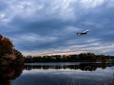 A large commercial airplane navigates through a cloudy sky, flying over a serene lake. The lake reflects the scattered clouds and the surrounding autumnal trees, which exhibit a range of colors from green to orange. The scene is tranquil with a hint of an impending change in weather.