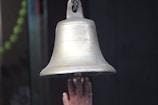 A hand reaches up to ring a large, metallic bell, which has inscriptions engraved on its surface. The hand is positioned below the bell, indicating an action of ringing it. Part of a green garland is visible in the background, suggesting a ceremonial or religious setting.