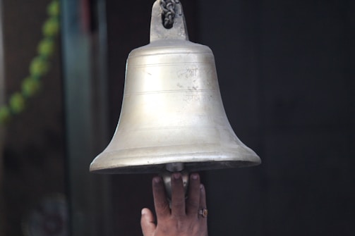 A hand reaches up to ring a large, metallic bell, which has inscriptions engraved on its surface. The hand is positioned below the bell, indicating an action of ringing it. Part of a green garland is visible in the background, suggesting a ceremonial or religious setting.