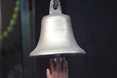 A hand reaches up to ring a large, metallic bell, which has inscriptions engraved on its surface. The hand is positioned below the bell, indicating an action of ringing it. Part of a green garland is visible in the background, suggesting a ceremonial or religious setting.