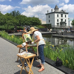 A woman is painting outdoors in a park setting. She is wearing a sunhat and standing in front of an easel with a canvas. There is a pond nearby with aquatic plants and yellow flowers. Trees and a historic-looking building are in the background, and there are people seated by the water.