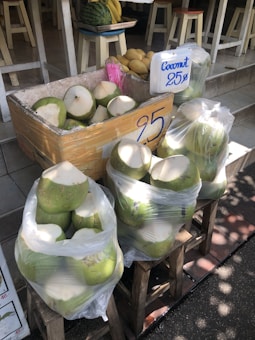A market stall displaying a collection of fresh coconuts in two clear plastic bags and an open crate. The coconuts are ready for drinking, with their tops cut off. A sign indicates a price of 25, possibly in a local currency. In the background, a tray holds a watermelon and bunches of bananas next to yellow mangoes, all placed on white stools near the entrance of a shop.