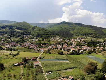 A small village nestled in a lush valley with numerous houses spread across the landscape. Surrounding the village are vast green fields and patches of agricultural land. In the background, there are rolling green hills and mountains under a partly cloudy sky.