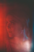 A woman is seen in a dimly lit environment with a soft focus effect. The lighting creates a warm red and cool blue hue, adding dramatic contrast. Her expression is contemplative, and she appears to be looking towards something.
