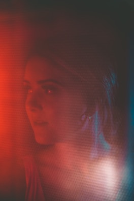 A woman is seen in a dimly lit environment with a soft focus effect. The lighting creates a warm red and cool blue hue, adding dramatic contrast. Her expression is contemplative, and she appears to be looking towards something.