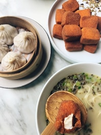 A bamboo steamer holds several steamed dumplings next to a bowl filled with a creamy noodle soup. Accompanying these is a plate with several fried snacks, one of which is on a wooden spoon ready for tasting.