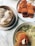 A bamboo steamer holds several steamed dumplings next to a bowl filled with a creamy noodle soup. Accompanying these is a plate with several fried snacks, one of which is on a wooden spoon ready for tasting.