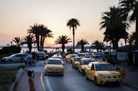 A serene sunset scene featuring palm trees along a coastal road. Several yellow taxis are parked, with people walking and sitting nearby. The sky is painted in shades of orange and pink as the sun sets behind the distant horizon.
