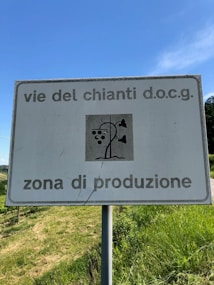 A sign with the text 'vie del chianti d.o.c.g. zona di produzione' against a clear blue sky. The sign includes an image of a stylized grapevine with grapes. Below the sign, there is a lush green field.