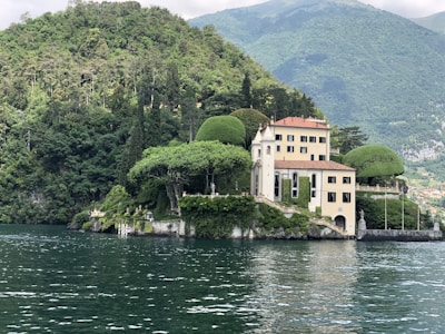A picturesque villa is nestled on the edge of a lush, forested hill overlooking a calm body of water. The building features a classic architectural style with light-colored walls and a terracotta roof. Surrounding the villa are meticulously maintained gardens and several sculpted trees. The backdrop consists of verdant hills and distant mountains under a partly cloudy sky.