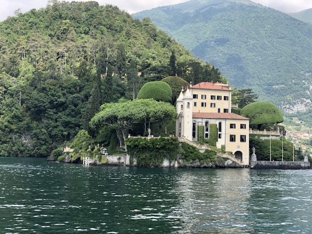 A picturesque villa is nestled on the edge of a lush, forested hill overlooking a calm body of water. The building features a classic architectural style with light-colored walls and a terracotta roof. Surrounding the villa are meticulously maintained gardens and several sculpted trees. The backdrop consists of verdant hills and distant mountains under a partly cloudy sky.