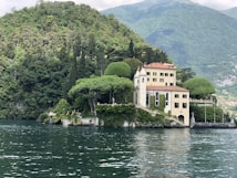 A picturesque villa is nestled on the edge of a lush, forested hill overlooking a calm body of water. The building features a classic architectural style with light-colored walls and a terracotta roof. Surrounding the villa are meticulously maintained gardens and several sculpted trees. The backdrop consists of verdant hills and distant mountains under a partly cloudy sky.