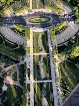 An aerial view of a landscaped park featuring large artistic depictions of hands on the grass. The scene includes pathways, trees, and people scattered around, with a road circling the area. The design of the park gives a sense of harmony and artistic expression within the urban environment.