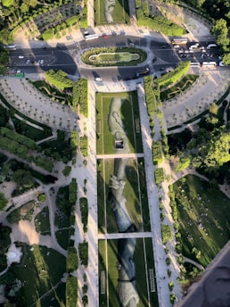 An aerial view of a landscaped park featuring large artistic depictions of hands on the grass. The scene includes pathways, trees, and people scattered around, with a road circling the area. The design of the park gives a sense of harmony and artistic expression within the urban environment.
