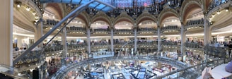 An opulent, multi-level shopping mall interior with ornate architectural details, featuring a large atrium and multiple floors with various stores. The space is bustling with shoppers amidst a grand setting, showcasing a blend of modern retail with classic design.