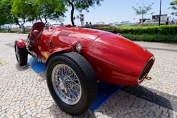A vintage red race car with sleek, polished curves is parked on a cobblestone path. The car's wire-spoke wheels and distinct front grille enhance its classic aesthetic. Lush green trees and a bright blue sky provide a vibrant background, while a few people and other vehicles are faintly visible in the vicinity.