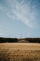 A wind turbine stands tall on a grassy hill under a partly cloudy sky. Power lines stretch across the scene, connecting to a distant pylon. The foreground features a golden field typical of harvested crops, surrounded by patches of green trees.