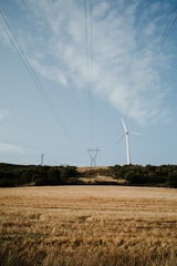 A wind turbine stands tall on a grassy hill under a partly cloudy sky. Power lines stretch across the scene, connecting to a distant pylon. The foreground features a golden field typical of harvested crops, surrounded by patches of green trees.